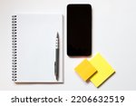 Top down flat lay view of notepad with white pages, pen, mobile phone and sticky note blocks on white background