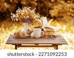 Bouquet of flowers, croissant, cup of tea or coffee, books on table in autumn garden. Rest in garden, reading books, breakfast, vacations in nature concept. Autumn time in garden on backyard