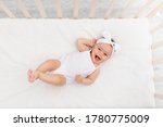 baby girl 6 months old lies in a crib in the nursery with white clothes on her back and laughs, looks at the camera, baby's morning, baby products concept