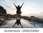 Silhouette of a man from the back sitting on a rock in the mountains, a joyful girl view from behind raised her hands up, reflection in a puddle of water on a stone, dawn in the mountains