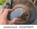 Small photo of car mechanic shows a worn brake pad, a worn brake shoe and a against the background of an automobile brake disc with signs of corrosion