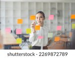 Small photo of Young Asian woman Creative team use post it notes to share idea sticky note on glass wall. Asian business people design planning and Brainstorming thinking sticky History notes concept.