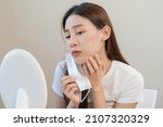 Small photo of Dermatology, puberty asian young woman, girl looking into mirror, allergy when wear mask and cosmetic, show squeezing pimple spot for removing from face.Beauty care from skin problem by acne treatment
