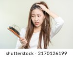 Serious, worried asian young woman, girl holding brush, show her comb, hairbrush with long loss hair problem after brushing, hair fall out on her hand in living room. Health care, beauty treatment.