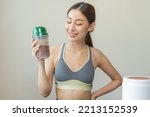 Diet meal replacement for weight loss, happy asian young woman, girl in sportswear, hand in holding protein shake bottle for drink supplement for muscle after workout at home. Healthy body care person