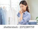 Small photo of Asian young housewife woman having bad smelling clothes hand holding breath nose with finger, sniff smelly dirty stinky musty, look disgusting from clothes after washed, laundry out of machine at home
