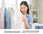 Small photo of Asian young housewife woman, girl expression face bad smelling clothes, sniff smelly dirty stinky musty, look disgusting from clothes after washed clothes, laundry out of machine in room at home.
