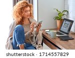 Young red-haired woman graphic designer working from home side hustle drinking hot coffee browsing internet on smartphone smiling curious