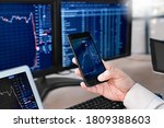 Man trader sitting at desk at office in front of monitors with stocks data holding smartphone browsing application monitoring price changes on candle bar online using digitla devices close-up