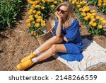 Beautiful young blond young woman in a blue dress wearing yellow rubber boots sitting with legs stretched in yellow bloom tulip field on a sunny summer day. Nature travel concept. Dutch countryside.