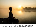 Silhouette of a person sitting on the pier against the sunset sky. Loneliness concept. The female looking at the rising sun over the lake in the fog. Reflection of the sun in the water surface.