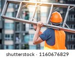 Young Asian maintenance worker man with orange safety helmet and vest carrying aluminium step ladder at construction site. Civil engineering, Architecture builder and building service concepts