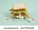 Small photo of Parts of a doll's body in a sandwich with salad and soft bread on a minimal background color. pop fun and quirky cannibalism