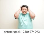 Small photo of Portrait of young Asian man wearing t-shirt rending his hair, screaming with close eyes and wide open mouth, holding hands on head. Isolated image on white background. People's emotion concept