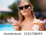 Cute woman with duckface applies sunscreen solar cream on face near swimming pool. Smiling pretty girl puts suntan cream from plastic bottle on skin in spa. Female in bikini with suntan lotion.