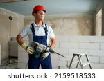 Professional construction worker in uniform standing with rotary hammer drill. Portrait of contractor in hardhat and overalls posing with jackhammer near step ladder and masonry indoors.