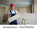 Small photo of Bricklayer or mason constructs wall of autoclaved aerated concrete blocks. Portrait of brickwork worker contractor standing with quality foamed concrete doing masonry on construction site.