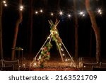 Small photo of Festive string lights illumination on boho tipi arch decor on outdoor wedding ceremony venue in pine forest at night. Vintage string lights bulb garlands shining above chairs at summer rural wedding.
