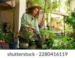 Young woman gardener in straw hat holding hand shovel taking care of potted plants. Junior caucasian female smiles standing in her little garden planting flowers in pots. Gardening and farming concept