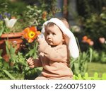 Small photo of Beautiful caucasian baby girl with a headband bunny ears on her head gives chocolate easter eggs in a shiny wrapper holding in a pen and sitting sideways on the lawn in the yard of the house on a clea
