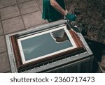 Small photo of One young Caucasian unrecognizable girl in green shorts and gloves paints a mirror frame with white paint with a brush, standing in the backyard of the house on a summer day, reflected in it, top vie