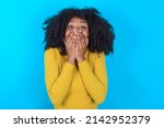 Small photo of Vivacious Young woman with afro hairstyle wearing yellow turtleneck over blue background , giggles joyfully, covers mouth, has natural laughter, hears positive story or funny anecdote