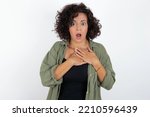 Scared young beautiful woman with curly short hair wearing green overshirt over white wall looks with frightened expression, keeps hands on chest, being puzzled to notice something strange, People.
