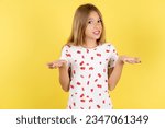 Small photo of Clueless beautiful caucasian kid girl wearing polka dot shirt shrugs shoulders with hesitation, faces doubtful situation, spreads palms, Hard decision