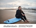 Dreamy body positive surfer woman looking away, while sitting with surf board