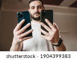 Portrait of a young bearded European man comparing two smartphones blue and gray. Technology, 2020, 2021, phone, new, choice. Holds in his hands, style, youth, latest technology, gadget, smart watch.