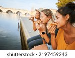 Three happy women sitting on the lakeside of the city eating pizza in a street stall. The happy girls enjoy the weekend together outdoors.