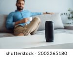 Young man sitting on sofa at smart home listening small portable wireless speaker. Focus on virtual voice digital sound assistant on table. Indian businessman using mini bluetooth stereo gadget 