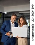 Small photo of Vertical portrait of mature Latin business man and European business woman discussing project on laptop in office. Two diverse confident professional partners colleagues business people work together.