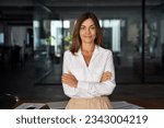Beautiful hispanic senior business woman with crossed arms smiling at camera. European or latin confident mature good looking middle age leader female businesswoman on office background, copy space.