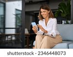 European businesswoman CEO holding smartphone using fintech application sitting on sofa in modern office. Smiling Latin Hispanic mature adult professional business woman using mobile phone cellphone. 