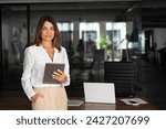 Small photo of Latin Hispanic mature adult professional business woman looking at camera and smiling. European businesswoman CEO holding digital tablet using fintech tab application standing at workplace in office.