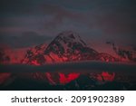Small photo of Surreal mountain landscape with great snowy mountains lit by dawn sun among low clouds. Fantasy alpine scenery with high mountain pinnacle at sunset or at sunrise. Big glacier on top in neon red light