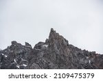 Gloomy mountain landscape with high sharp rockies with snow in gray rainy sky. Closeup of sharp rocks and peaked top in gray cloudy sky. Bleak overcast scenery with mountain range with pointy peak.