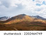 Small photo of Scenic motley autumn landscape with sunlit mountain top under gray dramatic cloudy sky. Vivid autumn colors in mountains. Multicolor high hill in sunlight with shadows of clouds in changeable weather.