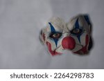 The scary face of a clown looking into the camera through a hole in the partition. Close-up. The face of a man wearing a creepy clown mask peeking at his victim through the hole. A creepy clown maniac