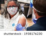 Small photo of San Diego, CA USA - Mar 17 2021: Woman wearing a mask gets vaccinated in her left arm for covid 19 at a gymnasium in San Diego by a firefighter wearing a face shield