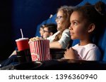 Beautiful african girl with funny hairstyle watching excited movie in cinema. Wonderful little child sitting with friends, eating popcorn and smiling. Concept of entertainment and enjoyment.
