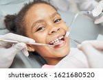 Charming little afro american girl sitting in dental chair, smiling and looking at camera during medical treatment at modern clinic. Concept of health care ad pediatrics