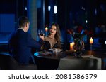 Charming young couple in elegant clothes toasting with glasses of red wine while sitting at table in dark atmosphere. Romantic date at home. Happy relationship.