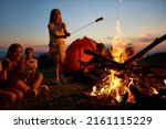 Happy teen girl frying sausage over fire, having fun at campsite at dusk. Side view of group of kids sitting around big campfire, roasting meal at twilight against colorful sky. Concept of camping.
