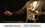 Small photo of Male orchestra conductor wearing tux standing in front of music stand, controlling musicians by moving his hands and baton. Studio shot on black background