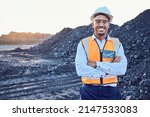 A young African mine worker wearing protective wear is looking at the camera with coal mine in the background