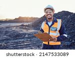 A young African mine worker wearing protective wear is looking at the camera while holding a clipboard with coal mine in the background