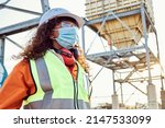 Small photo of A young African woman mine worker wearing protective wear is looking off camera while wearing a covid 19 face mask with coal mine equipment in the background
