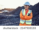 Small photo of A young African woman mine worker wearing protective wear is looking off camera with coal mine equipment in the background